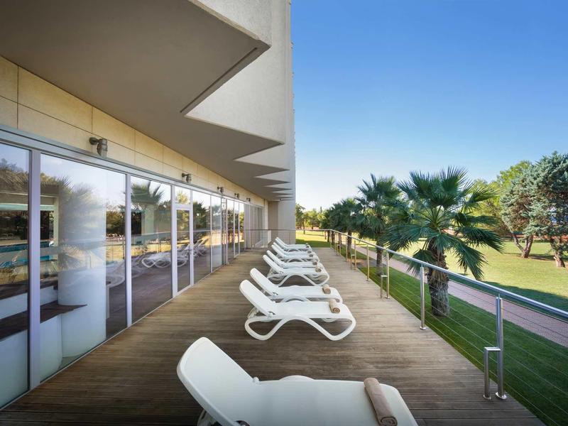 Balkon mit weißen Liegestühlen, Glasgeländer, Holzterrasse und Blick auf grüne Wiese und Palmen.