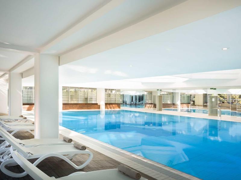Helles Hallenbad mit blauem Wasser, weißen Liegestühlen und großen Fenstern für Tageslicht.