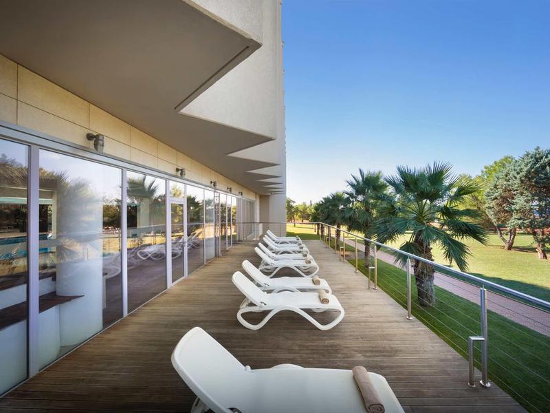 Balkon mit Liegestühlen, Glasbrüstung, Palmen und Rasenfläche unter klarem blauem Himmel.