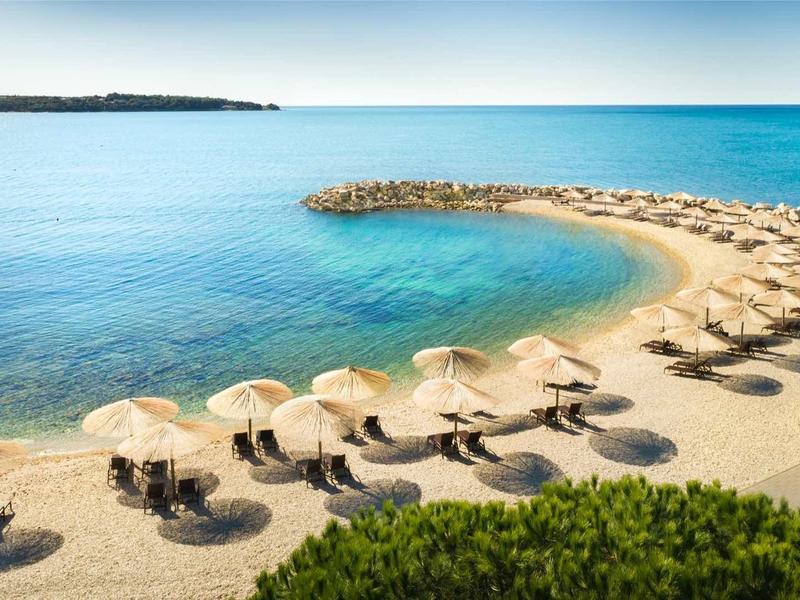 Kleiner Sandstrand mit vielen Liegen und Strohm Sonnenschirmen am ruhigen blauen Meer.