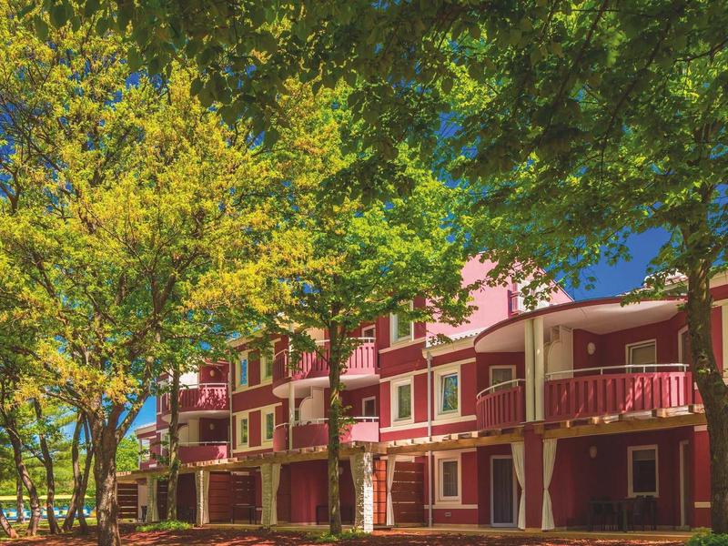 Wielopiętrowy budynek z czerwonymi balkonami otoczony zielonymi drzewami pod czystym niebem