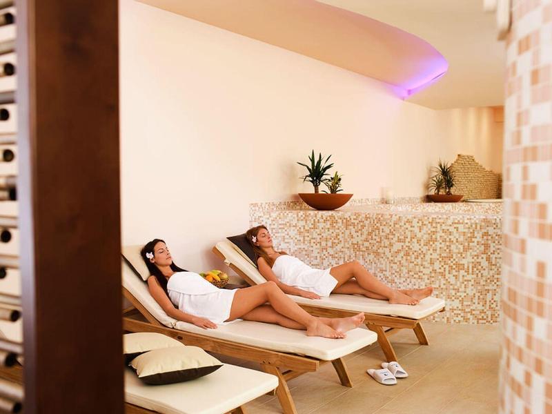 Zwei Frauen entspannen auf Liegen in einem hellen Wellnessraum mit warmem Licht und Pflanzen.