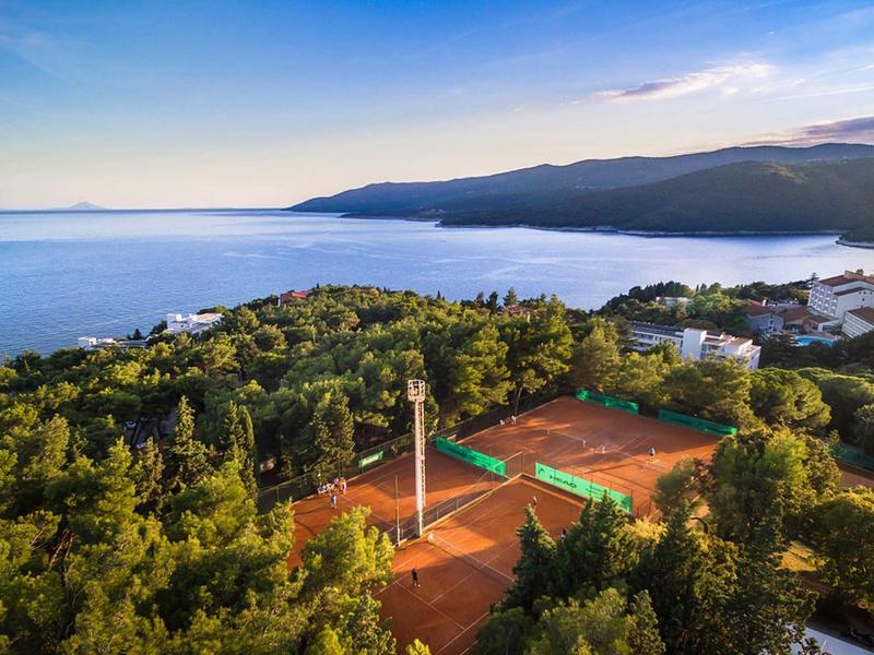 Blick auf Tennisplätze in grüner Küstenlandschaft mit Meer und Bergen im Hintergrund bei Sonnenuntergang.