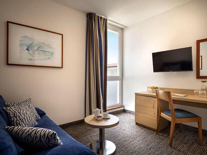 Kleiner Raum mit blauem Sofa, rundem Tisch, Schreibtisch, Stuhl, Wandbild und TV an der Wand.