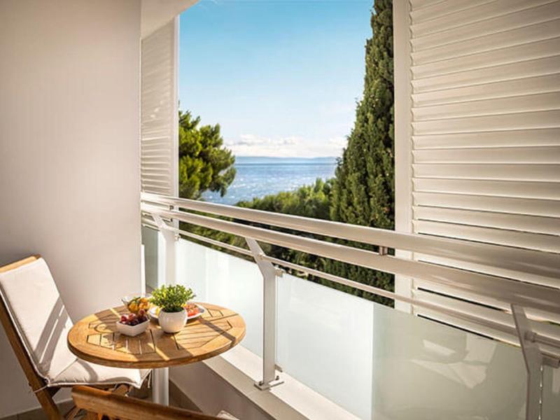 Kleiner Balkon mit Holzstuhl, rundem Tisch und Blick auf Meer und grüne Bäume.