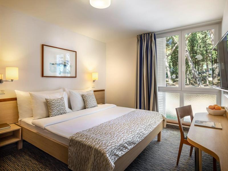 Gemütliches Hotelzimmer mit Doppelbett, Schreibtisch, Stuhl und Fenster mit Blick auf Bäume.