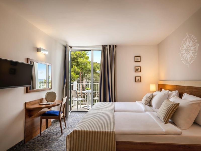 Helles Hotelzimmer mit Doppelbett, Schreibtisch, Fernseher und Balkon mit Blick ins Grüne.