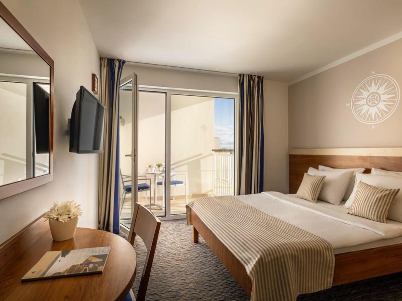 Helles Hotelzimmer mit Doppelbett, Schreibtisch, TV und Balkon mit Meerblick.