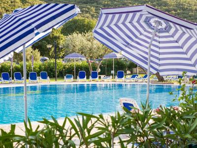Poolbereich mit blau-weißen Sonnenschirmen und Liegestühlen vor grünen Hügeln.