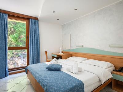 Hell eingerichtetes Hotelzimmer mit Doppelbett, blauen Vorhängen und Fenster zur begrünten Außenseite.