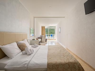Helles Hotelzimmer mit Doppelbett, Schreibtisch, Fernseher und Blick auf einen Balkon mit Sitzgelegenheiten.
