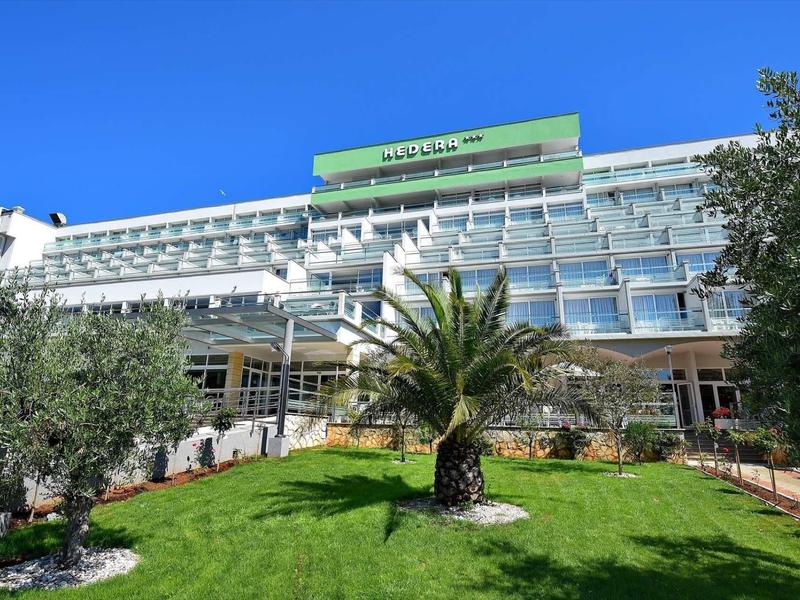 Modern hotel building with glass facade and manicured garden with palm trees under clear blue sky.