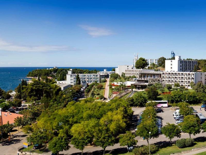 Blick auf ein Küstenhotel mit großem Parkplatz, viel Grün und blauem Meer im Hintergrund.