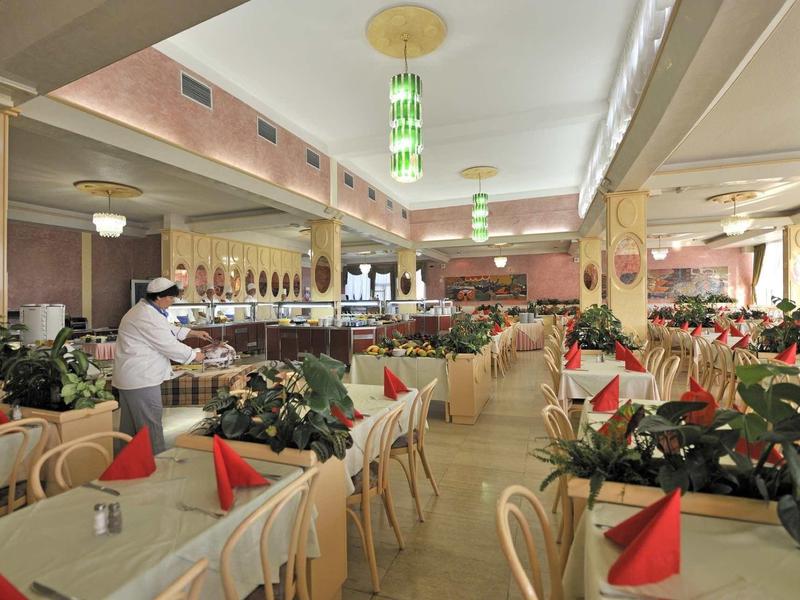 Großer, heller Restaurantraum mit Holzstühlen, Tischen und roten Servietten, Dekorationspflanzen.