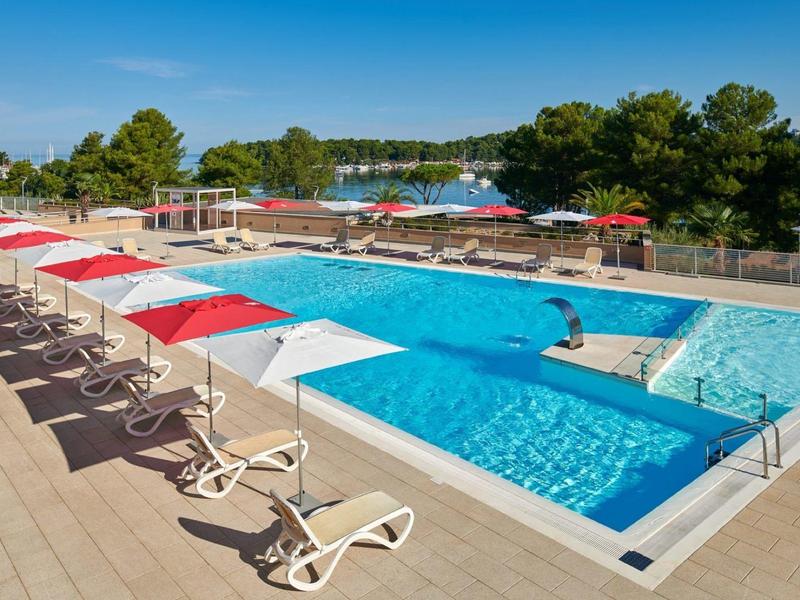 Piscina all'aperto con sedie a sdraio e ombrelloni rossi e bianchi accanto agli alberi sotto un cielo limpido.