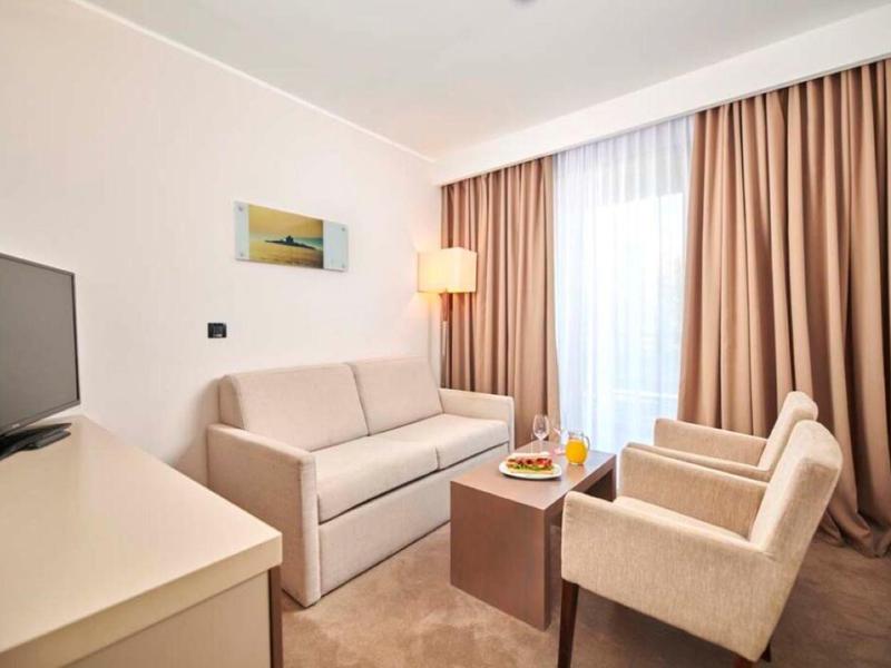 Camera d'hotel luminosa con divano, poltrone, TV e un tavolo davanti a grandi tende.