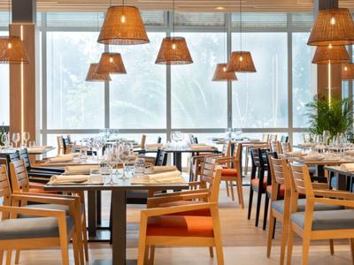 Bright restaurant room with wooden tables, chairs, and large lamps in front of windows showing snow outside.