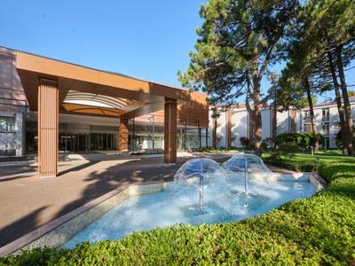 Modern hotel with water fountain, surrounded by green bushes and tall trees on a sunny day.