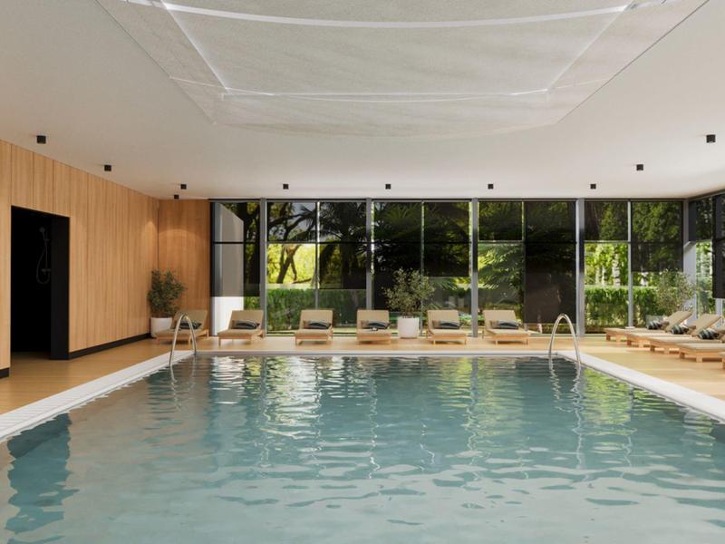 Vnitřní bazén s velkými okny, dřevěným obložením a lehátky obklopený zelení.