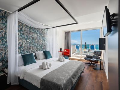 Modernes Hotelzimmer mit großem Bett, Sessel und Blick auf Meer und Balkon.