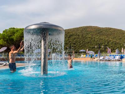 Ein Pool mit Springbrunnen und spielenden Personen vor bewaldeten Hügeln und Sonnenschirmen.