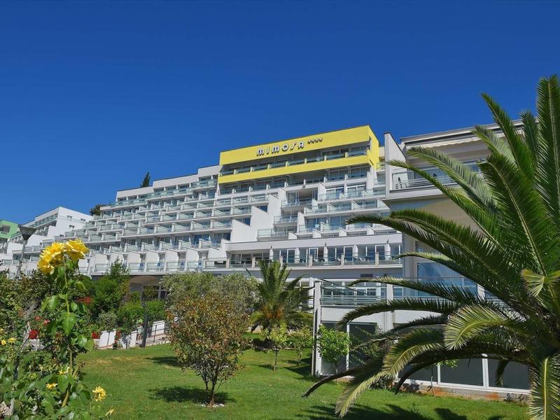 Modern hotel complex with multiple balconies, surrounded by palm trees and flowering plants under clear sky.