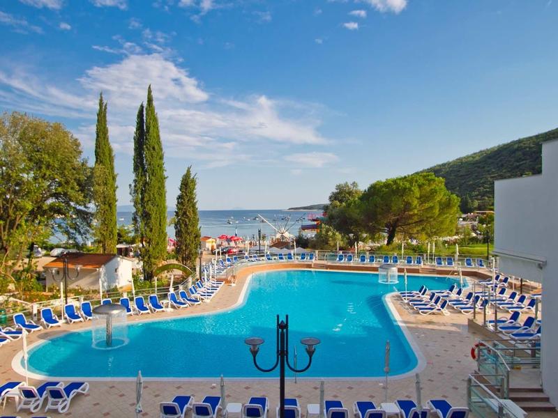 Large outdoor pool with lounge chairs, trees, and sea view in the background.