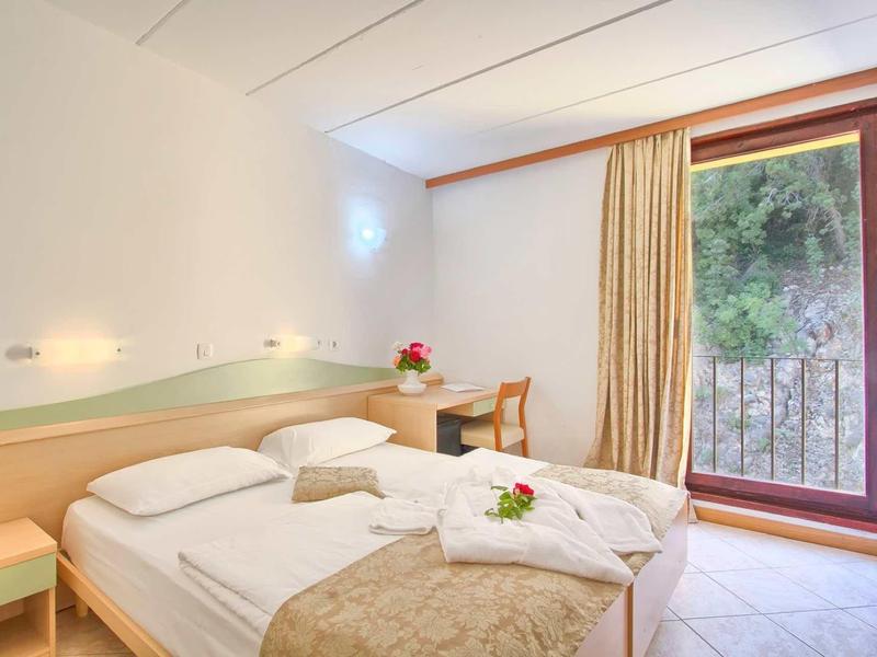 Bright hotel room with double bed, desk, and balcony overlooking greenery.