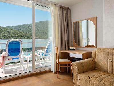 Habitación de hotel con balcón, zona de estar, escritorio y vistas a colinas boscosas y agua.