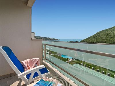 Balcón con silla azul y barandilla de vidrio con vista al mar y colinas verdes