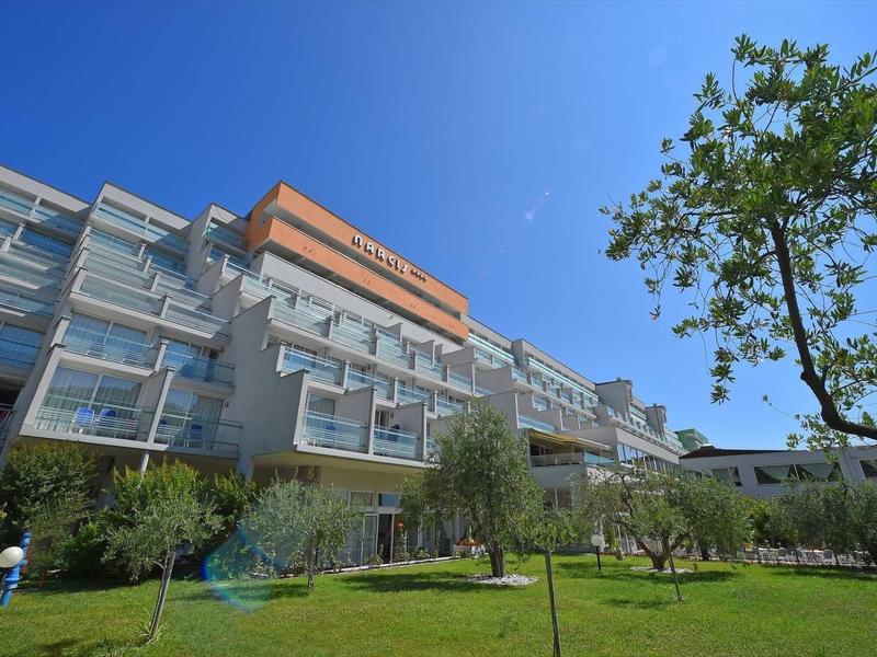 Modern hotel building with balconies, surrounded by green lawn and trees under a blue sky.