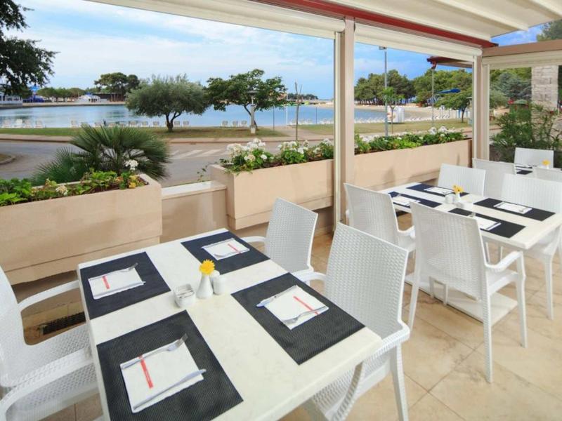 Cafe terrace with white tables and chairs overlooking a seaside promenade and the ocean.