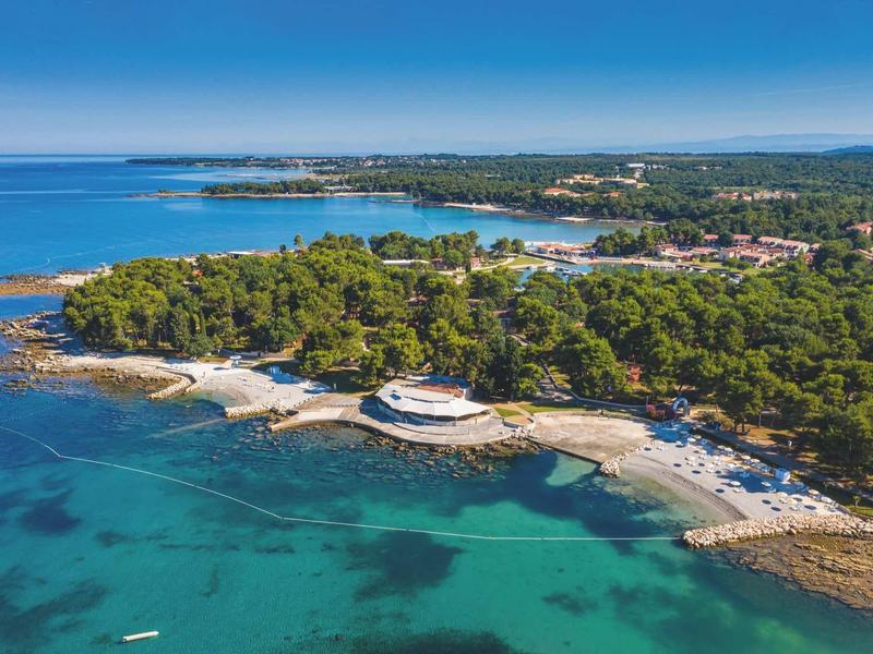 Aerial view of a coastline with clear water, forest, and a round building on the beach.