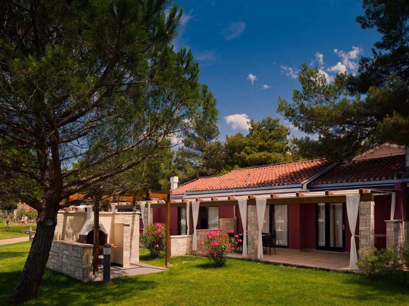 A holiday home with a red tiled roof surrounded by greenery and trees under a blue sky.