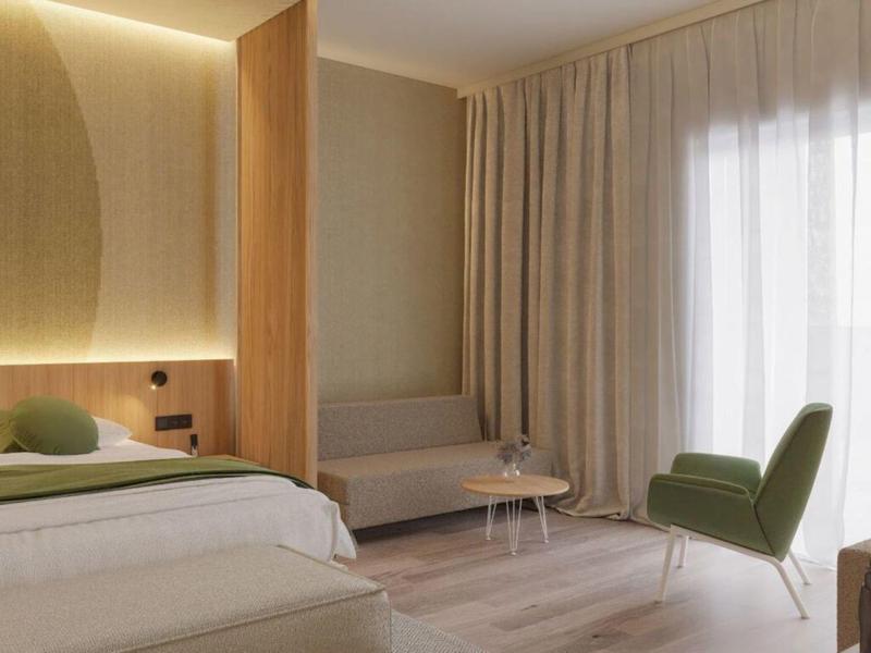Habitación de hotel moderna con cama, sillón verde y zona de estar a la luz del día.