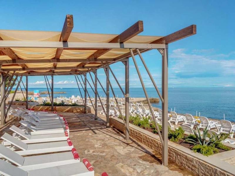 Strand mit weißen Liegen unter Holz-Pergola, blauer Himmel, Meer und Felsen im Hintergrund.
