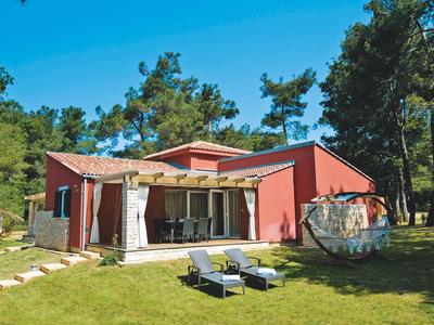 Modern rood huis met terras en ligstoelen in een groene tuin onder een blauwe lucht.