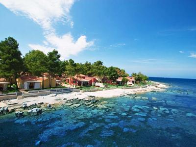 Kustlijn met helder water, bomen en kleine huizen onder een blauwe lucht.