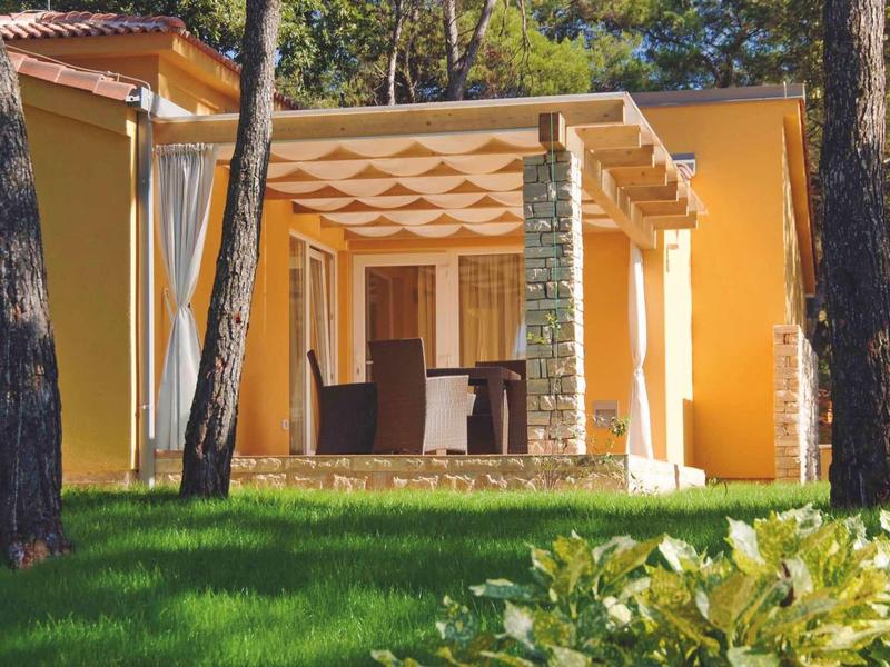 Geel vakantiehuis met terras en pergola omgeven door groen en bomen.