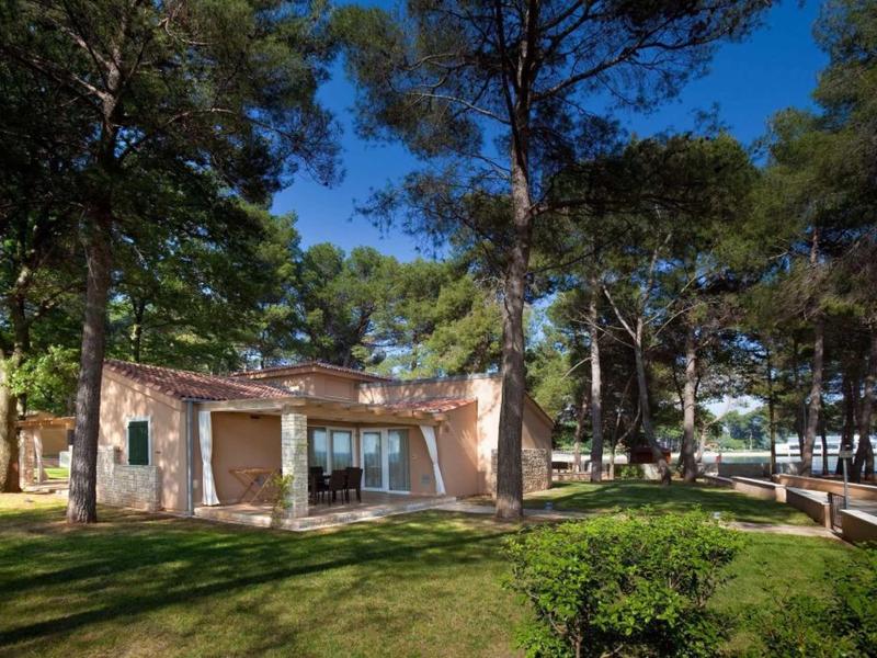 Een klein vakantiehuis met een terras, omringd door bomen en groen gras onder een blauwe lucht.