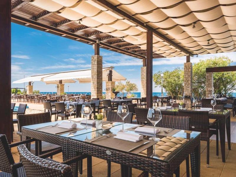 Open hotelrestaurant met gedekte tafels en uitzicht op zee.