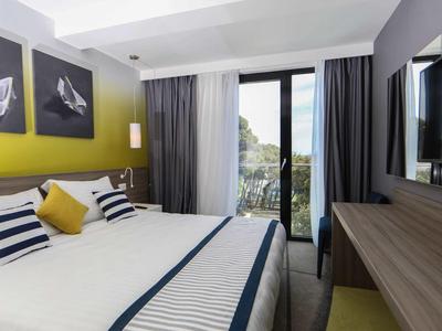 Modern hotelkamer met groot bed, gele accentmuur en uitzicht op een terras.