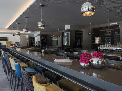Moderne hotelbar met lange tafels, kleurrijke stoelen en hangende bolverlichting.