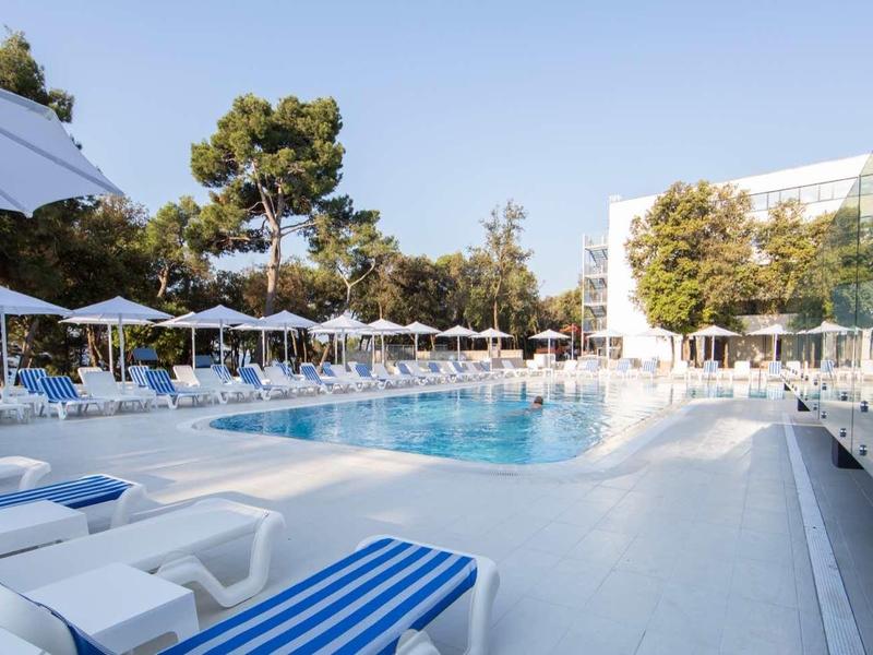 Nowoczesny basen hotelowy z leżakami i parasolami pod drzewami w pogodny dzień.