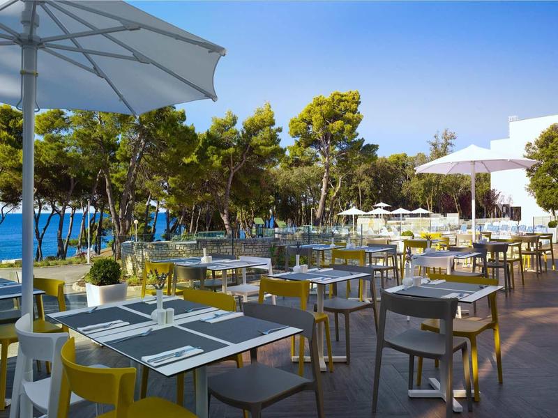 Restauracja na zewnątrz z białymi stołami, żółtymi krzesłami i parasolami przy sosnach nad morzem.