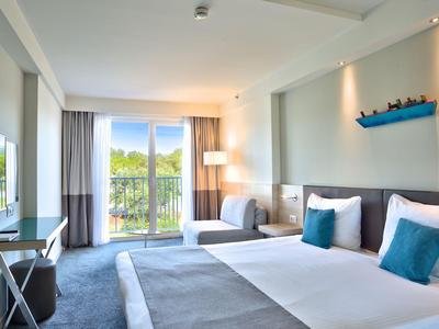 Modernes Hotelzimmer mit Doppelbett, Sofa, Fernseher und Balkon mit Blick auf Bäume.