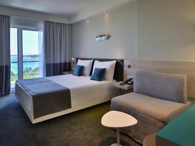 Nowoczesny pokój hotelowy z podwójnym łóżkiem, sofą, stołem i widokiem na morze przez duże okna.