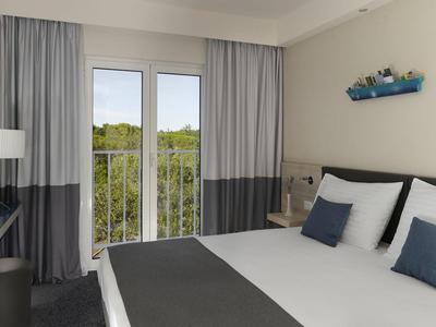 Modernes Hotelzimmer mit Doppelbett, Schreibtisch und Balkon mit Blick auf grüne Bäume.