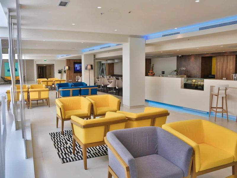 Nowoczesny hotelowy salon z żółtymi fotelami, szarą sofą i barem w tle.