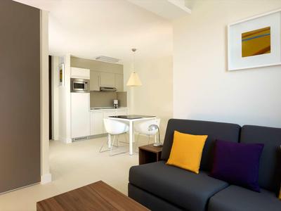 Nowoczesny salon z sofą, małym stołem jadalnym i otwartą kuchnią w neutralnych kolorach.